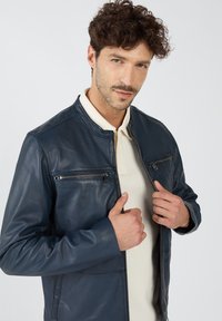 Marineblaue Lederjacke mit Stehkragen, mit Reißverschlüssen versehenen Brusttaschen und langen Ärmeln. GlatteTextur und taillierte Passform.