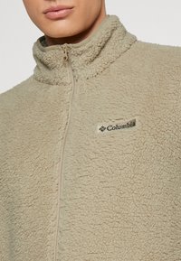 Beżowa polarowa kurtka z wysokim kołnierzem, pół-zamkiem z przodu oraz detalem logo Columbia. Materiał jest puszysty i teksturowany, zapewniając ciepło.
