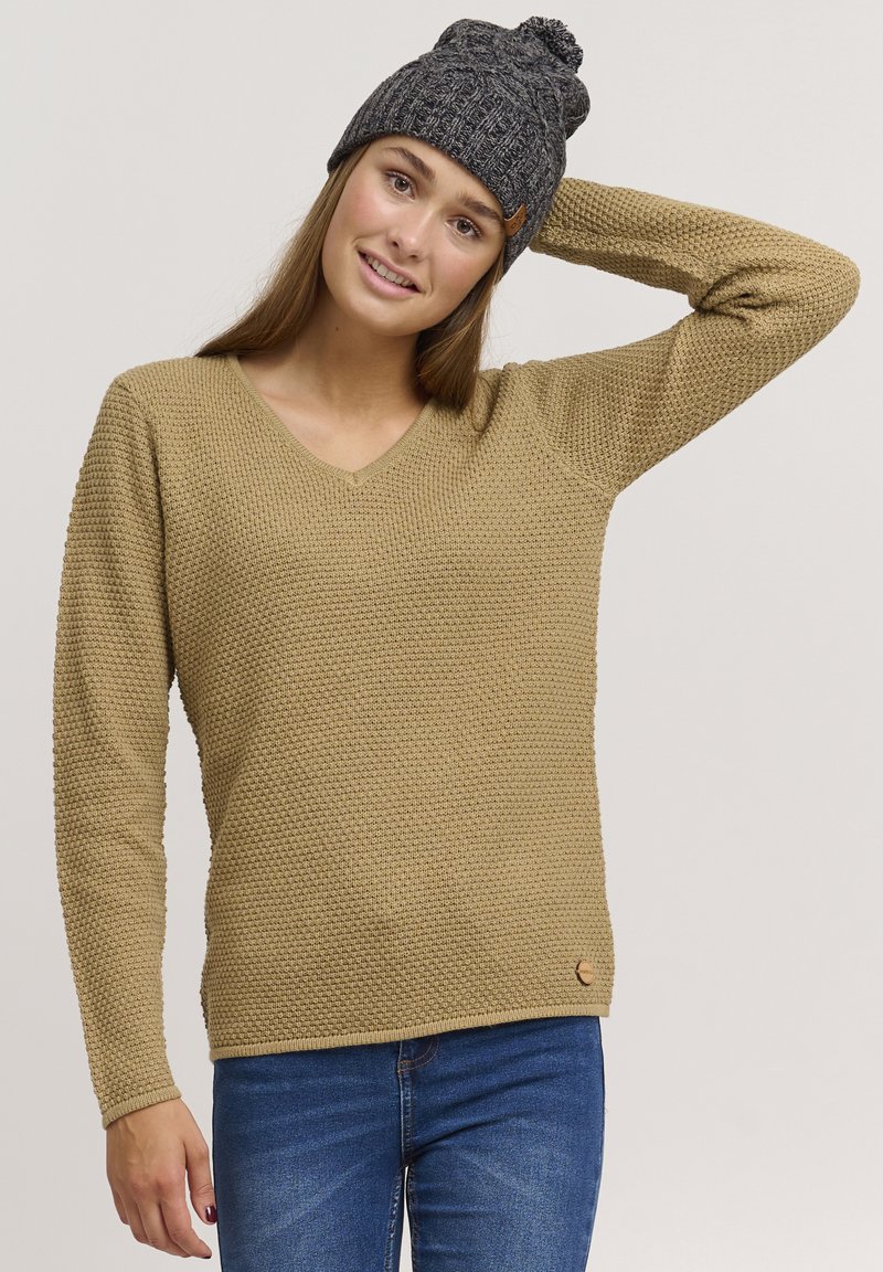 Maglione beige lavorato a maglia con scollatura a V e motivo testurizzato, abbinato a un berretto grigio lavorato a maglia con pom-pom in cima.