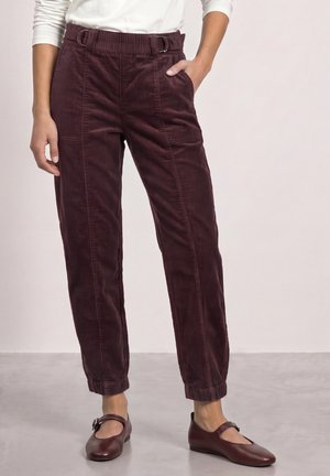 Femme portant un pantalon en velours côtelé bordeaux foncé taille haute avec des revers aux chevilles et des chaussures plates Mary Jane assorties, debout sur un sol gris clair.