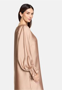 Langes lichtbraunes Satin-Kleid mit lockerer Passform, das über Puffärmel und geraffte Manschetten verfügt. Glatte Textur und minimalistisches Design.