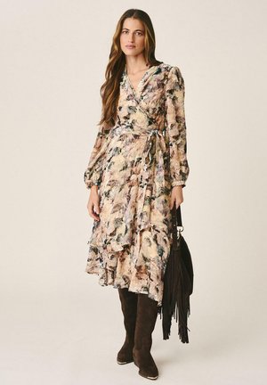 Femme aux longs cheveux portant une robe portefeuille fleurie et des bottes marron montantes, tenant un sac frangé noir sur un fond uni.