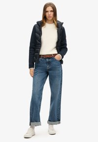 Chaleco acolchado azul marino sobre un suéter crema, combinado con unos vaqueros de pierna ancha de color azul claro y zapatillas blancas. Un cinturón trenzado marrón acentúa el conjunto.