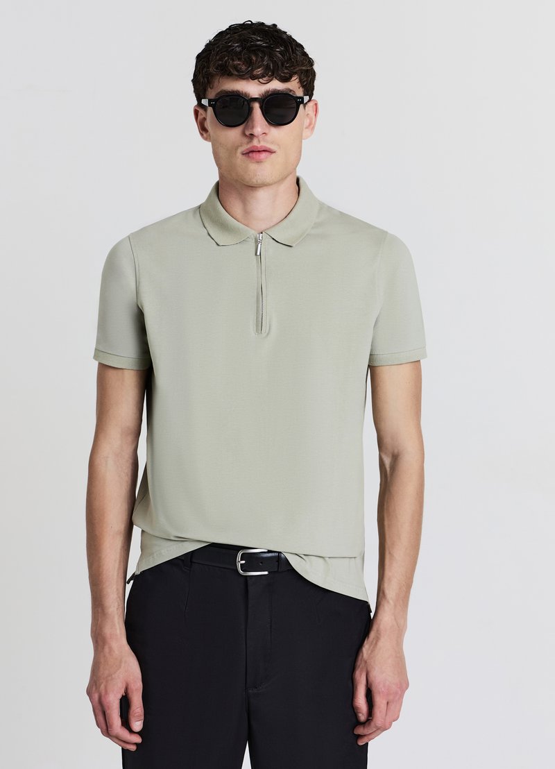 Polo léger vert clair à manches courtes avec un col zippé, associé à un pantalon noir. Le tissu semble lisse avec un léger éclat.