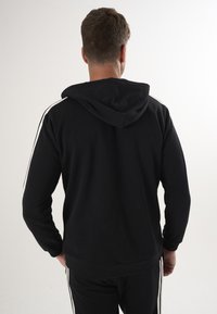 Schwarzer Kapuzenpullover mit langen Ärmeln, ausgestattet mit weißen Seitenstreifen und einem weichen, strukturierten Stoff. Ansicht von hinten.
