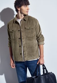Olivově zelená kordurová košile s knoflíkovým zapínáním, dvěma náprsními kapsami a ohrnutými rukávy, v kombinaci s modrými džíny a černou koženou taškou.