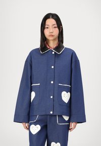 Blue denim jacket featuring a black leather collar, white button closures, and heart-shaped crochet accents on pockets. Boxy fit with long sleeves.
