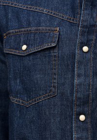Nahaufnahme eines dunkelblauen Jeanshemds mit orangefarbener Naht, Klappentasche mit Druckknopf und vorderen Druckknopfverschlüssen.