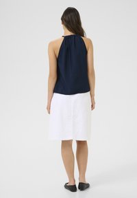 Haut bleu marine à col halter en tissu texturé, associé à une jupe blanche longueur genou. Le modèle porte des chaussures plates noires.