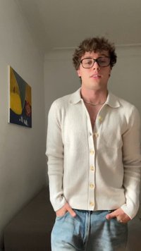 Joven con pelo rizado y gafas, vistiendo un cárdigan blanco de botones y vaqueros azules, de pie en interiores cerca de una pintura mural abstracta colorida.
