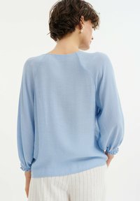 Hellbluse mit langen Ärmeln, gerafften Bündchen und lockerer Passform. Aus weichem, strukturiertem Stoff hergestellt. Die Rückansicht zeigt einen kurvigen Ausschnitt.