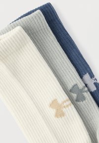 Τρία διπλωμένα κάλτσες με ριμπ πλεκτή υφή σε λευκό, γκρι και ναυτικό μπλε, κάθε ένα με ένα μικρό, αντίθετο λογότυπο Under Armour κοντά στο τελείωμα.