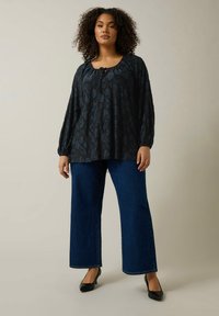 Schwarz gemusterte Bluse mit Rundhalsausschnitt und langen Ärmeln, kombiniert mit hochgeschnittenen blauen Schlagjeans und schwarzen High Heels.