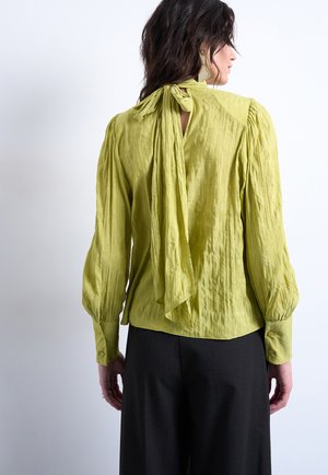 Femme portant un chemisier texturé vert citron à manches longues avec un nœud noué au cou, associé à un pantalon noir à jambes larges, tournée dos à nous.