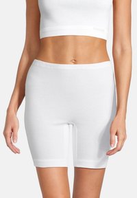 Kvinde iført hvide, tætsiddende shorts til midt på låret og en hvid cropped tanktop, vist fra midt på torsoen til midt på låret mod en ensfarvet baggrund.