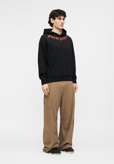 Jaunas vyras, dėvintis juodą gobtuvo megztinį su raudonu užrašu „Palm Angels“ ir laisvas rudas kelnes, stovintis priešais baltą foną.