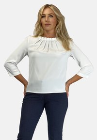 Biała bluzka z plisowanym dekoltem, rękawami o długości 3/4, gładkiego materiału i prostym kroju, w zestawieniu z granatowymi spodniami w wzory.