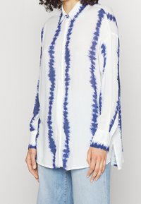 Camicetta bianca con strisce blu tie-dye, caratterizzata da maniche lunghe, colletto e fessure laterali. Realizzata in un tessuto leggero e morbido.