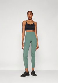 Schwarzer Sport-BH und hoch taillierte grüne Leggings. Glatter, enganliegender Stoff mit matter Oberfläche. Modell trägt schwarze Sportschuhe.