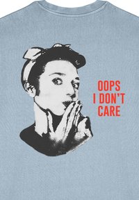 T-shirt bleu clair avec un graphique noir et blanc d'une personne portant un bandeau, les mains aux lèvres. Le texte indique "OUPS JE M'EN FOUS" en rouge.