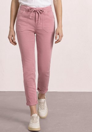 Mujer con pantalones rosa de talle alto con bolsillos delanteros y cinturón atado, combinados con zapatillas blancas, caminando sobre un suelo gris claro.