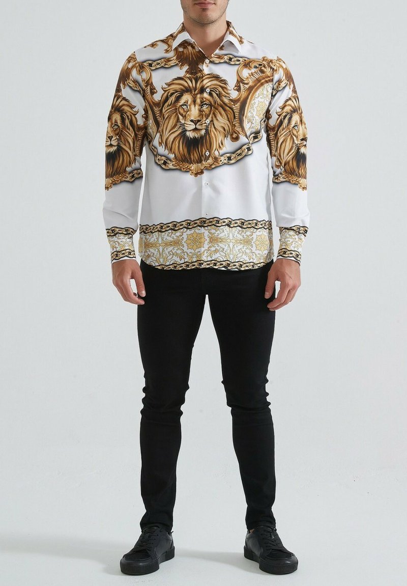 Chemise blanche à manches longues ornée de grandes impressions de lions dorés et de détails ornementaux, complétée par une bande dorée et noire à motifs au niveau de l'ourlet.