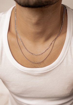 Silver layered necklaces with different chain styles rest on a bare chest, paired with a white ribbed tank top collar.