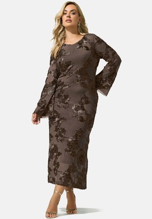 Femme portant une longue robe marron avec une broderie florale en sequins et des manches longues évasées, associée à des talons dorés à bride à la cheville, se tenant avec assurance.
