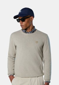 Maglione beige a maglia con collo rotondo, con un piccolo logo sul petto; indossato sopra una camicia a quadri blu e bianca; abbinato a un cappellino blu navy e occhiali da sole.