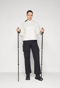 Giacca leggera bianca con zip e cappuccio, pantaloni cargo neri con tasche, scarpe da trekking nere, in mano due bastoni da trekking regolabili.