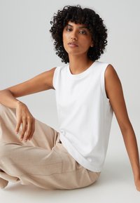 Weißes ärmelloses Oberteil aus Baumwolle mit rundem Ausschnitt, kombiniert mit beigen, strukturierten Weitbeinhosen. Einfaches und minimalistisches Design.