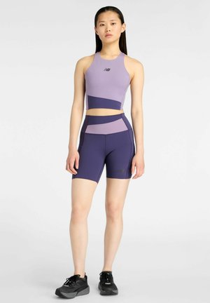 Ung kvinde iført lyserød og marineblå atletisk crop top og cykelshorts med sorte sneakers, stående på hvid baggrund.