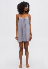 Γυναίκα με σγουρά μαλλιά που φοράει αμάνικο, άνετο φόρεμα με μπλε και λευκές ρίγες, όρθια ξυπόλυτη μπροστά σε απλό φόντο.