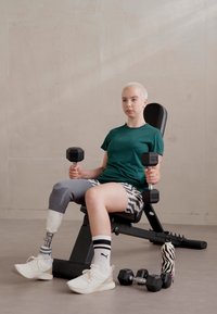 Dumbbell u rukama osobe u zelenoj majici i šarenim šortsevima, koja sjedi na crnoj klupi za vježbanje s plastičnom bocu za vodu sa zebrinim uzorkom u blizini.