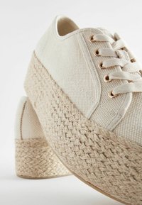 Beige canvas tennarit, joissa on kudottu juuttialusta. Ominaisuudet: pyöreät nauhat ja metalliset reikänauhat. Teksturoitu kangas, jossa on ommeltuja yksityiskohtia.