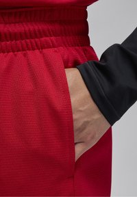 Punaiset urheilushortsit, joissa on teksturoidusta kankaasta valmistettu materiaali, joustava vyötärönauha ja sivutasku; yhdistetty mustaan pitkähihaiseen paitaan, jossa käsi taskussa.