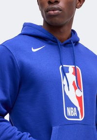 Άνδρας που φοράει μπλε φούτερ Nike με το λογότυπο του NBA στο στήθος, στραμμένος ελαφρώς προς τα αριστερά σε απλή φόντο.