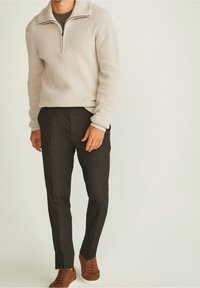 Lichtbeige gebreide trui met een ritskraag, gecombineerd met donkere tapered broek en bruine sneakers, wat een samenhangende, casual uitstraling creëert.