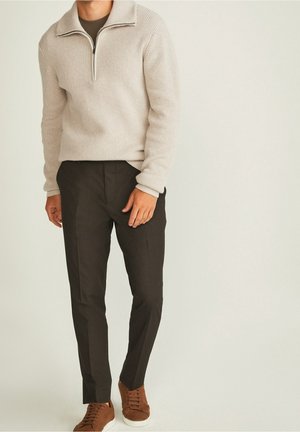 Pull léger beige en tricot avec un col zippé, associé à un pantalon sombre et ajusté ainsi qu'à des baskets marron, créant un look décontracté et cohérent.