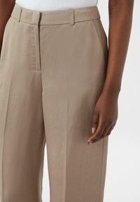 Beżowe spodnie o szerokich nogawkach z gładką teksturą, z przodu z fałdami, dopasowanym pasem i kieszeniami bocznymi. Ręka oparta na nogawce spodni.