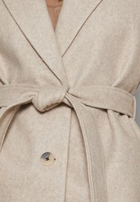Béžový kabát z vlněné směsi s klopovým límcem, samostatně vázaným páskem a velkým knoflíkem na přední straně, s měkkým, strukturovaným povrchem.