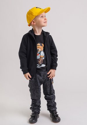Młody chłopiec w żółtej czapce, czarnej kurtce, koszulce z nadrukiem niedźwiedzia, ciemnych spodniach w wzory i czarnych trampkach patrzy w górę w prawo na jednolitym tle.