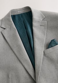 Blazer gris clair avec doublure à motif bleu sarcelle foncé et pochette assortie, présenté à plat sur un fond clair.