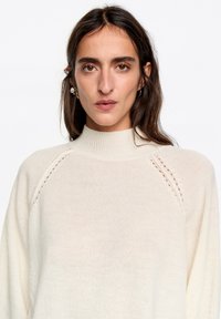 Kremowy sweter dziany z wysokim kołnierzem, prążkowanymi krawędziami i ozdobnym przeszyciem na ramionach; miękka tekstura i luźny krój.