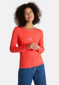 Frau mit lockigem Haar lächelt, trägt ein korallfarbenes langärmliges Shirt mit einem kleinen rosa Herzen auf der Brust und eine blaue Jeans.