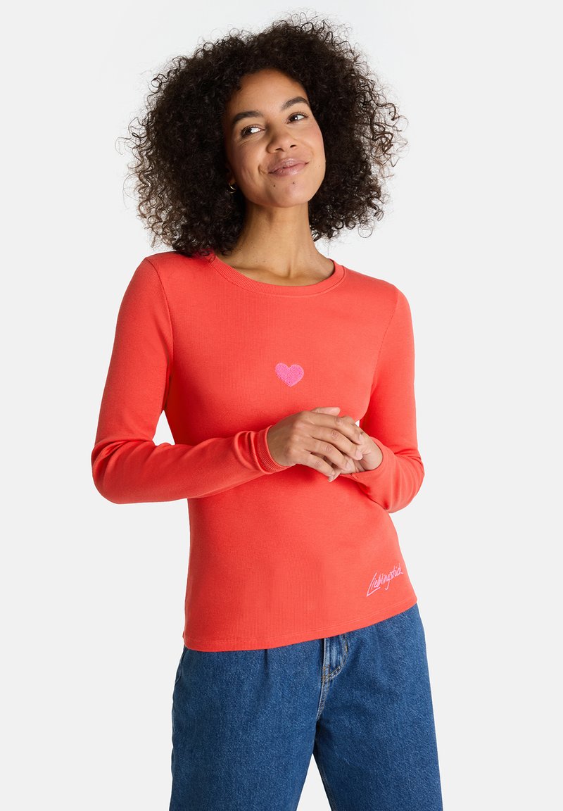Frau mit lockigem Haar lächelt, trägt ein korallfarbenes langärmliges Shirt mit einem kleinen rosa Herzen auf der Brust und eine blaue Jeans.
