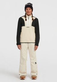 Crème en zwarte fleece jacket met hoge kraag, halve rits aan de voorkant en logo, gecombineerd met beige sneeuwbroeken en bruine sneeuwlaarzen. Zwarte gebreide beanie.