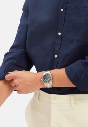 Mand ruller navyblå langærmet skjorte op, iført sølvur og cremefarvede bukser med knappet linning.