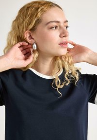 Marineblauwe t-shirt met een witte kraag, met een relaxte pasvorm en zachte textuur. Het model draagt ovale zilveren oorbellen en raakt haar haar aan.