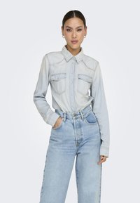 Camisa de mezclilla azul claro con cuello, botonadura frontal y dos bolsillos en el pecho. Combinada con unos vaqueros de tiro alto de lavado claro con un corte relajado.